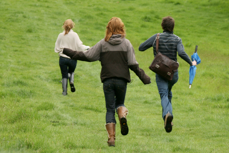 Three women with backs to us, one holding an umbrella, running down a grassy hill.