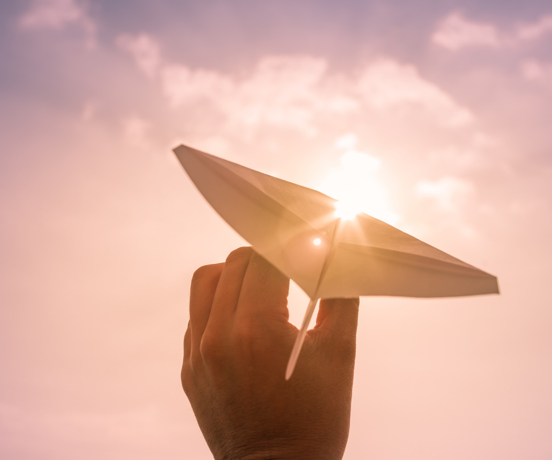 Hand holding a paper airplane with bright sun and clouds in the background.