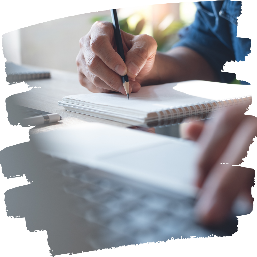 Close up of a man typing with one hand on a keyboard and taking notes on a pad with the other hand