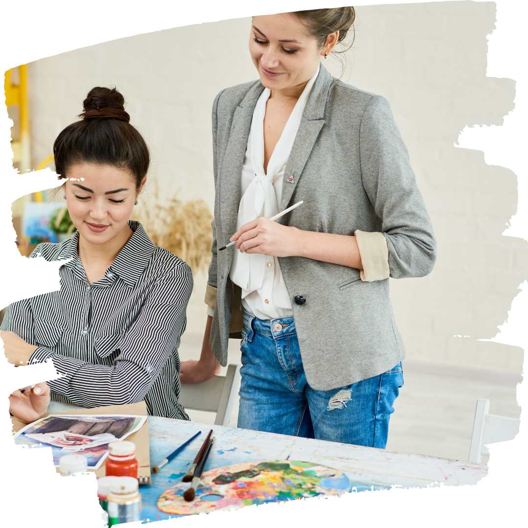 Woman standing with a paintbrush behind a seated woman with art supplies doing a painting