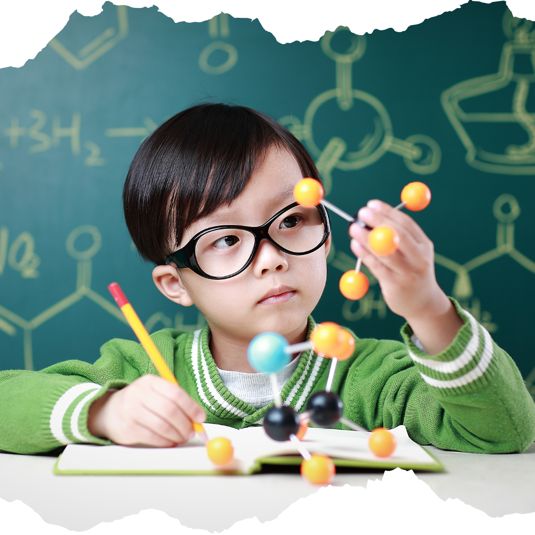 Young boy holding molecule models and writing on a notebook with a chalkboard behind him