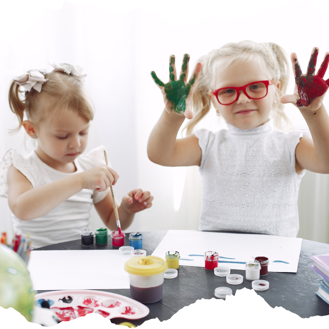 Two small girls finger painting at home, one is holding up her hands covered in red and green paint.